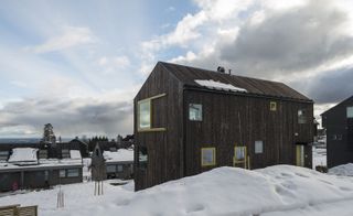 Alternative exterior view of the Oslo family house by STA during the day - the house features a dark wood exterior and windows and doors with yellow frames. There is snow on the ground and other houses nearby