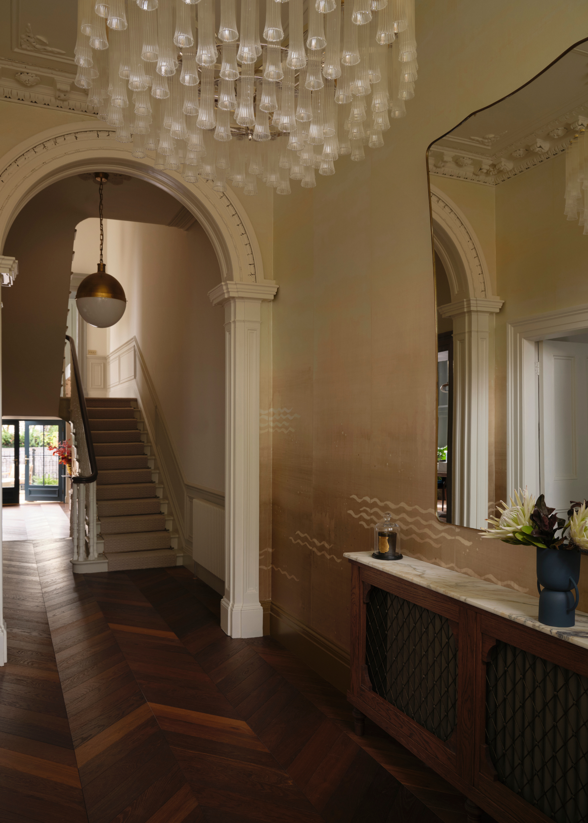 An entryway with herringbone wood flooring, a wood sideboard with a marble top, a vase of flowers, a candle, and a wall mirror beside a glass chandelier