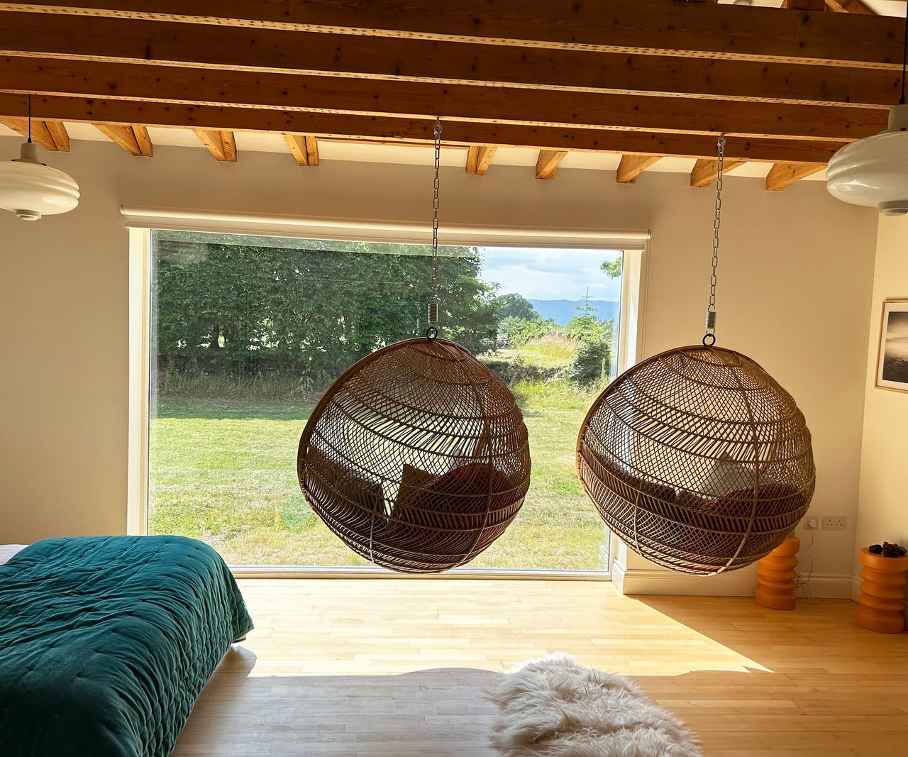 Two swinging egg chairs from a wooden beamed ceiling in a bedroom with views of the sea and garden through a floor to ceiling window