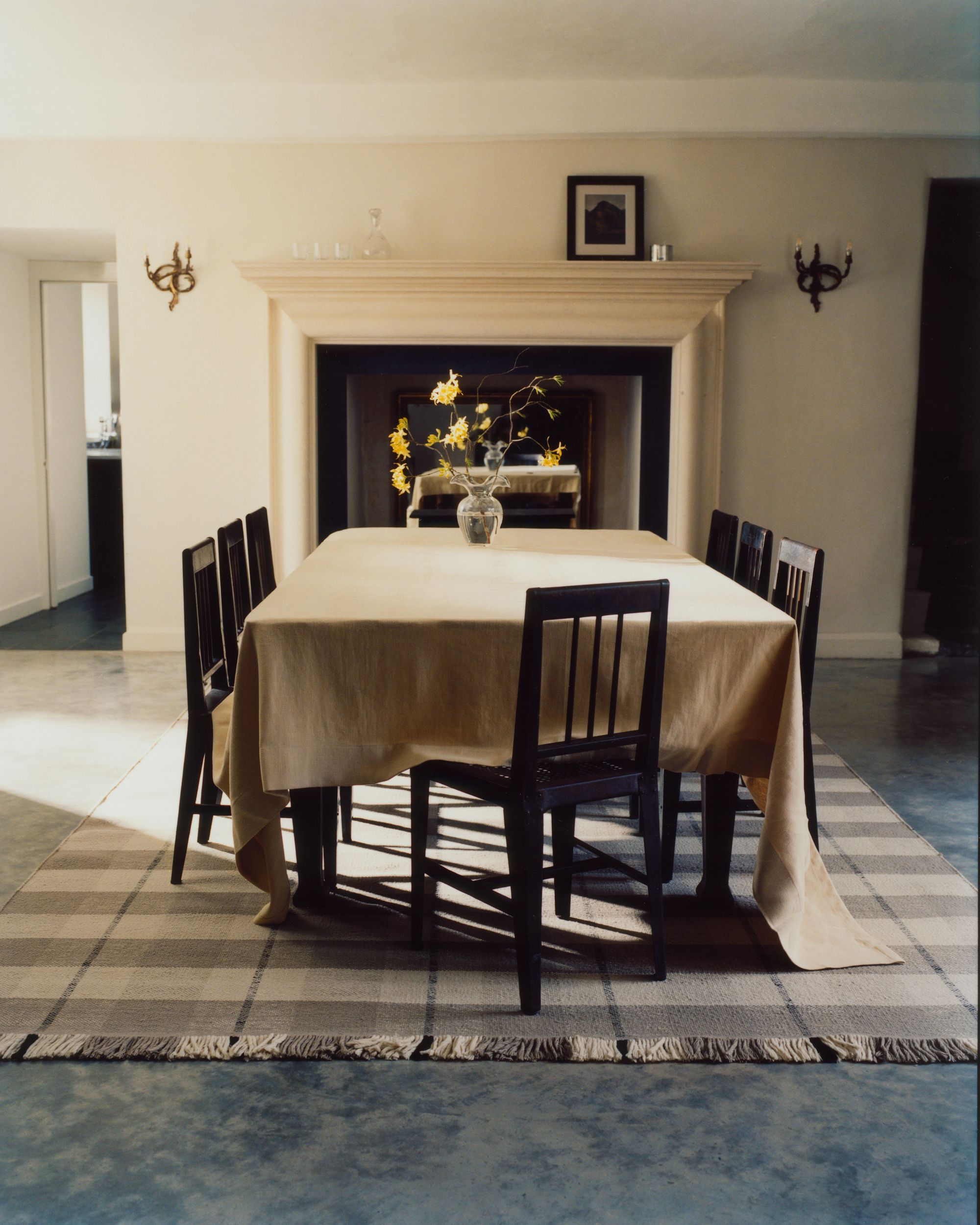 Image of a beige dining room with blue floors. There is a dining table in the middle with a beige tablecloth on it, black chairs around it, and a black and white plaid rug underneath.
