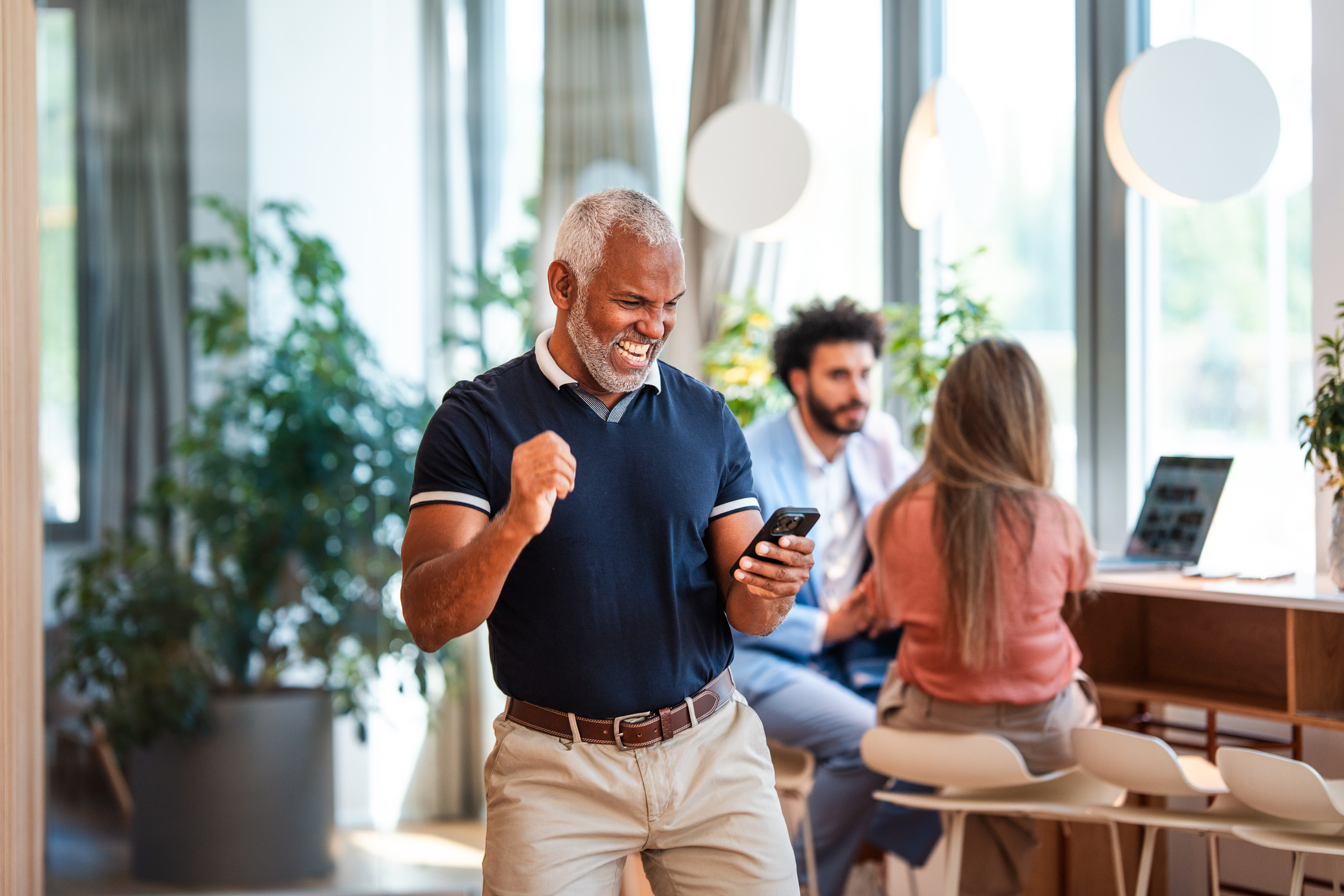 A mature Hispanic businessman is performing a victory dance with a smartphone in hand in a modern office featuring bright natural lighting and contemporary furnishings. Colleagues in the background are working on laptops, adding to the vibrant workplace atmosphere.