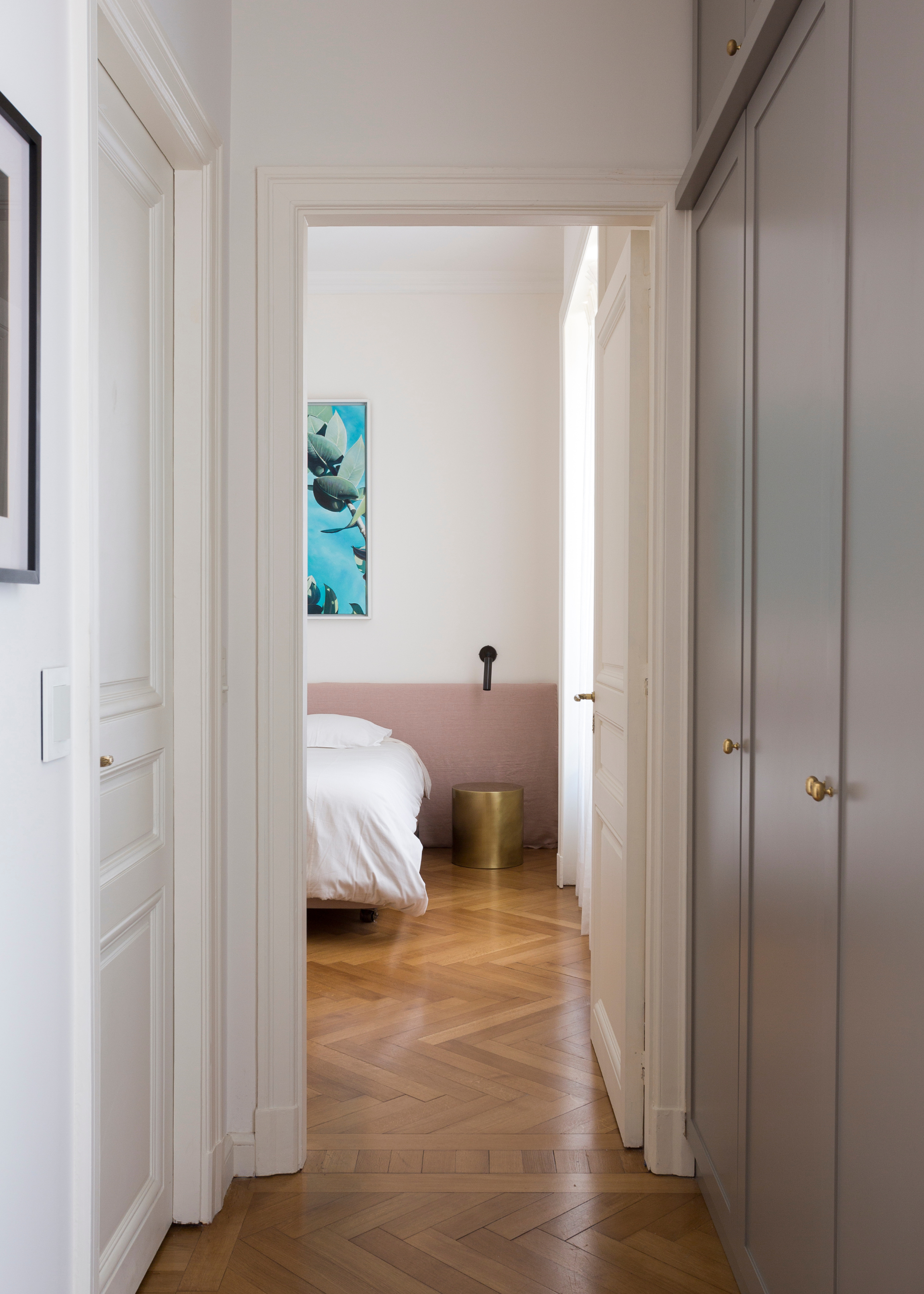 A corridor leading to a white bedroom with a colorful painting, and a brushed gold side stool