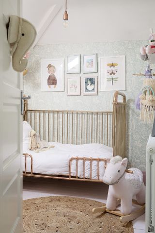 Nursery with pretty pastel wallpaper, gallery wall, gold cot bed and jute rug on floor with pink rocking horse