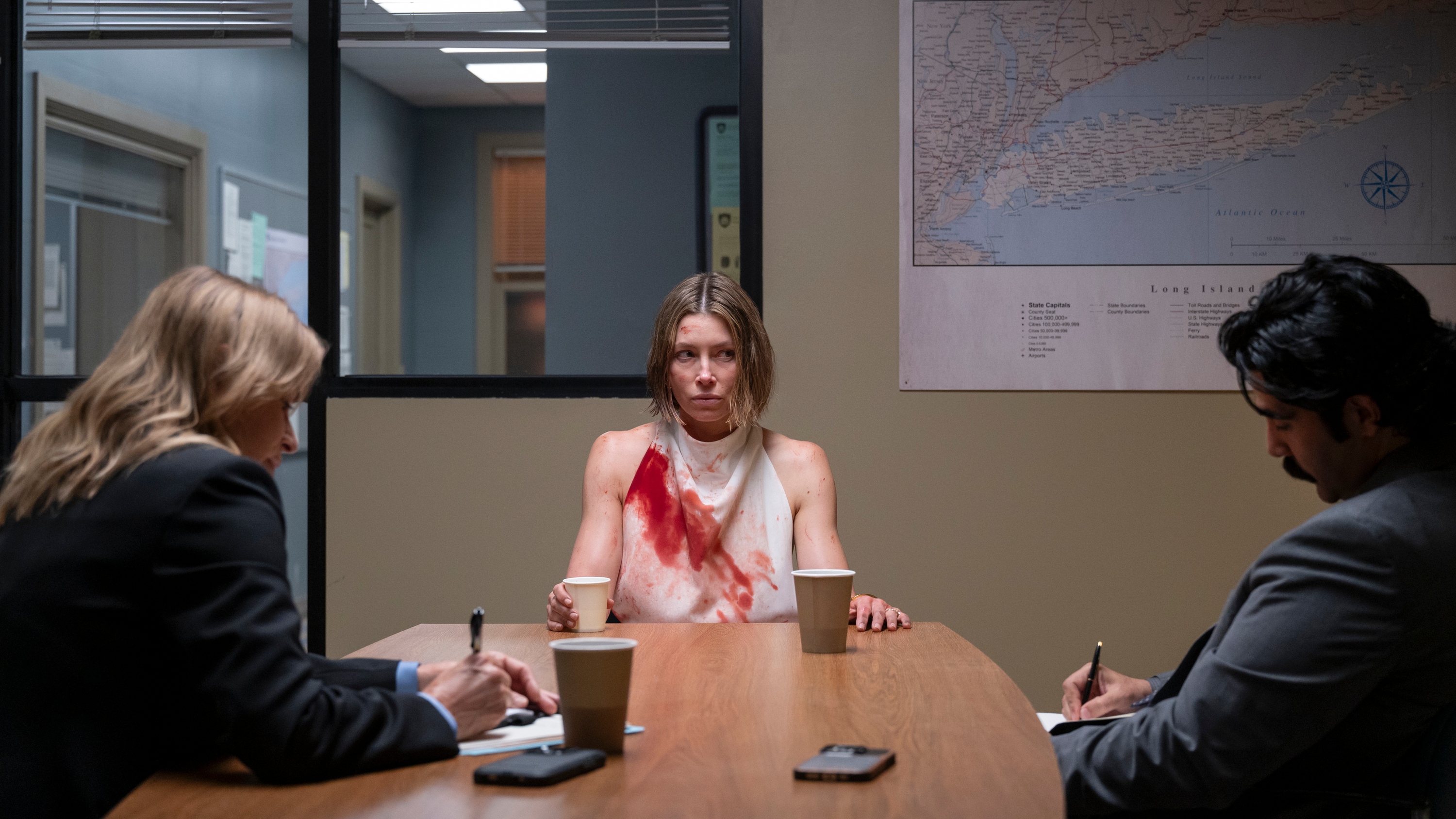 Police question Chloe in an interview room, she is looking upset and wearing a white dress which is covered in blood 