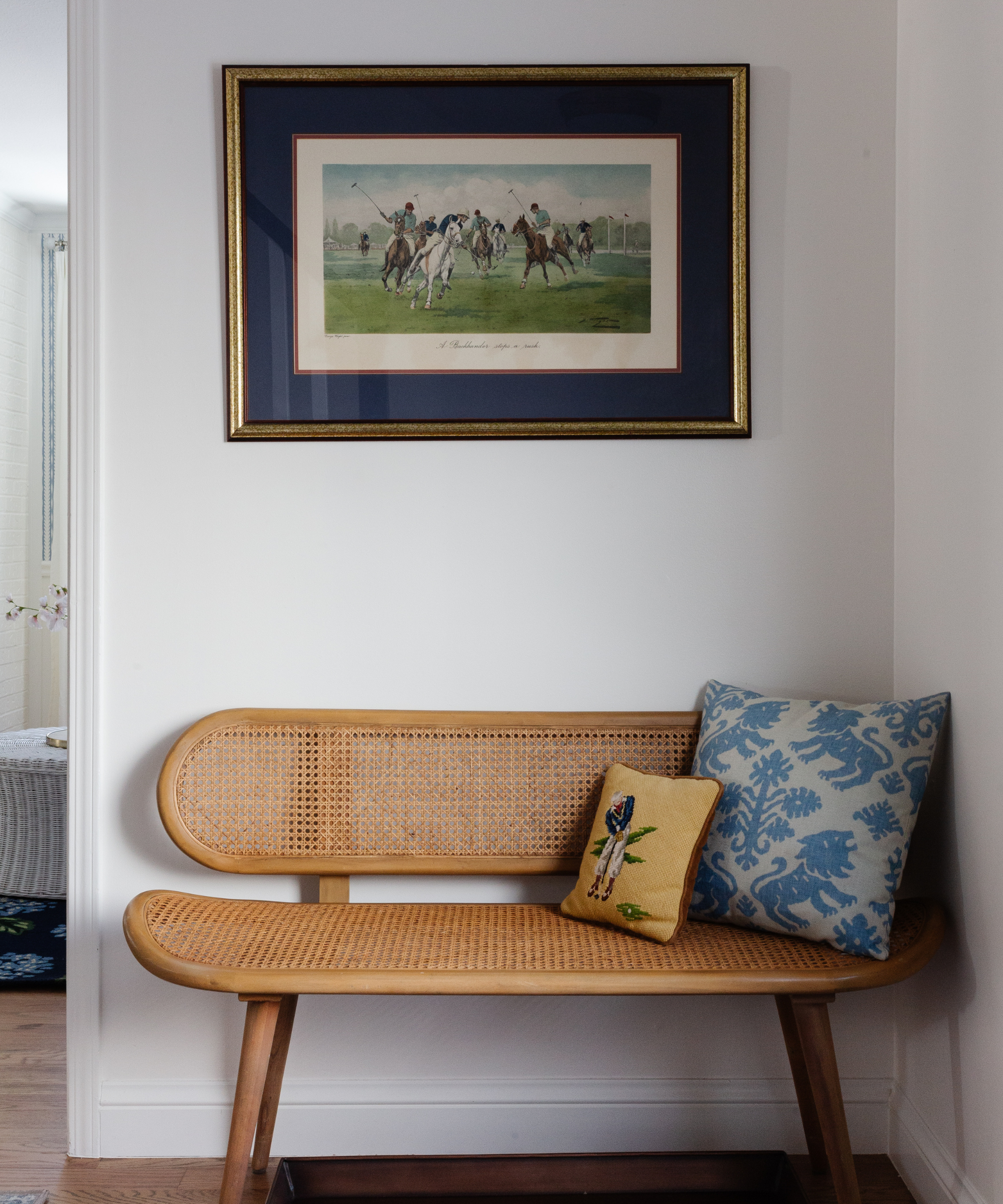 an entryway sitting area with a rattan bench with two pillows below a framed picture of people playing polo on horses