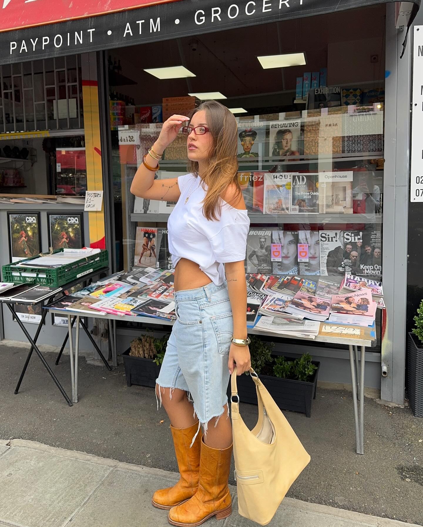 Fashion person Olivia Herring wears beige Frye boots, denim jorts a white of the shoulder top and is carrying a butter yellow bag. She is stood in front of a news agent in a recent image taken from her Instagram.