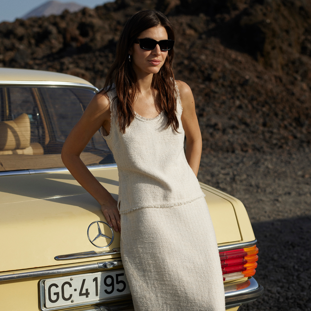 Model leaning on a car wearing a white dress and sunglasses sold at Whistles