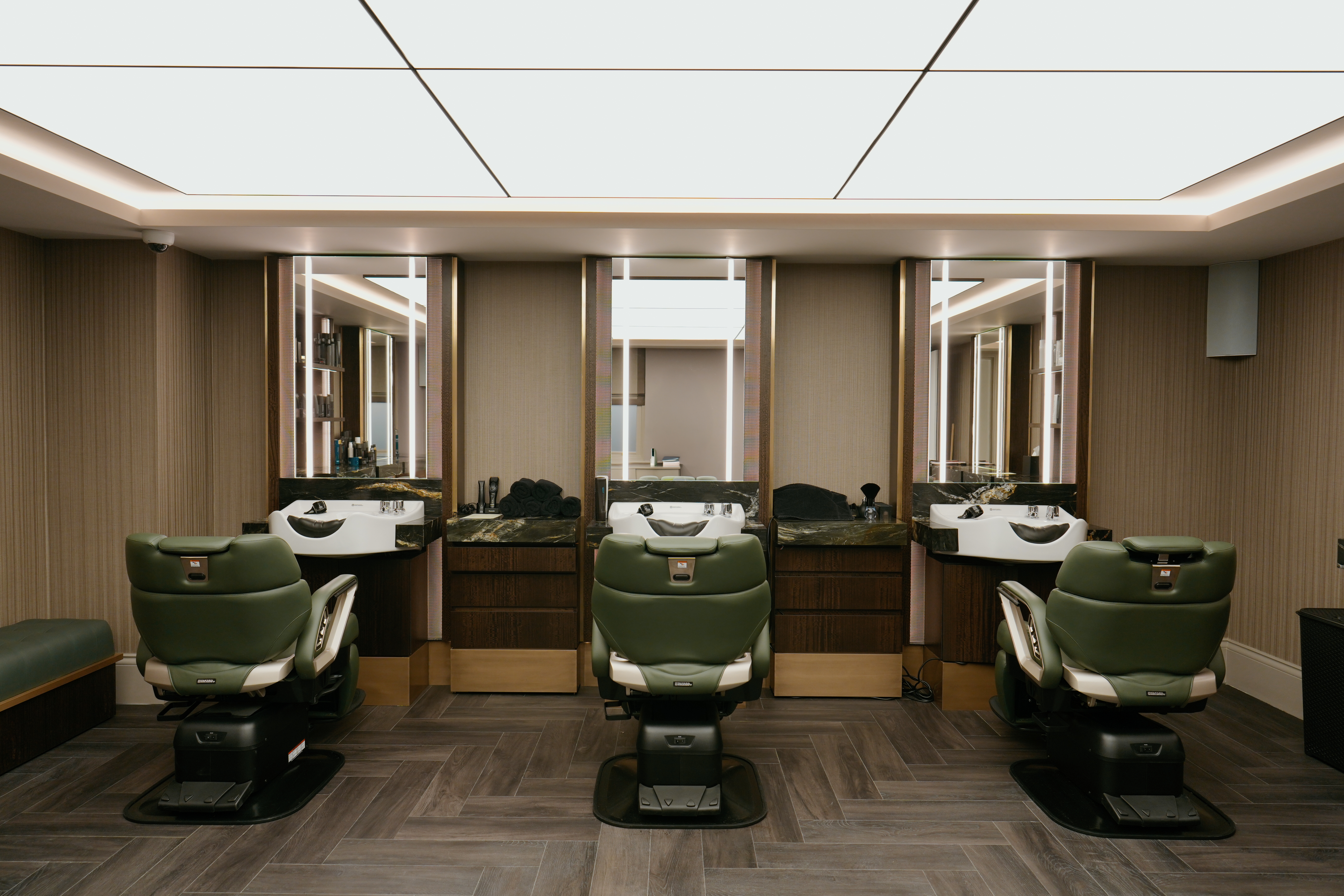 A retro-inspired barber shop features back-lit, coffered ceilings, wooden floors, and leather green hairdressing stations against a beige and brass color scheme.