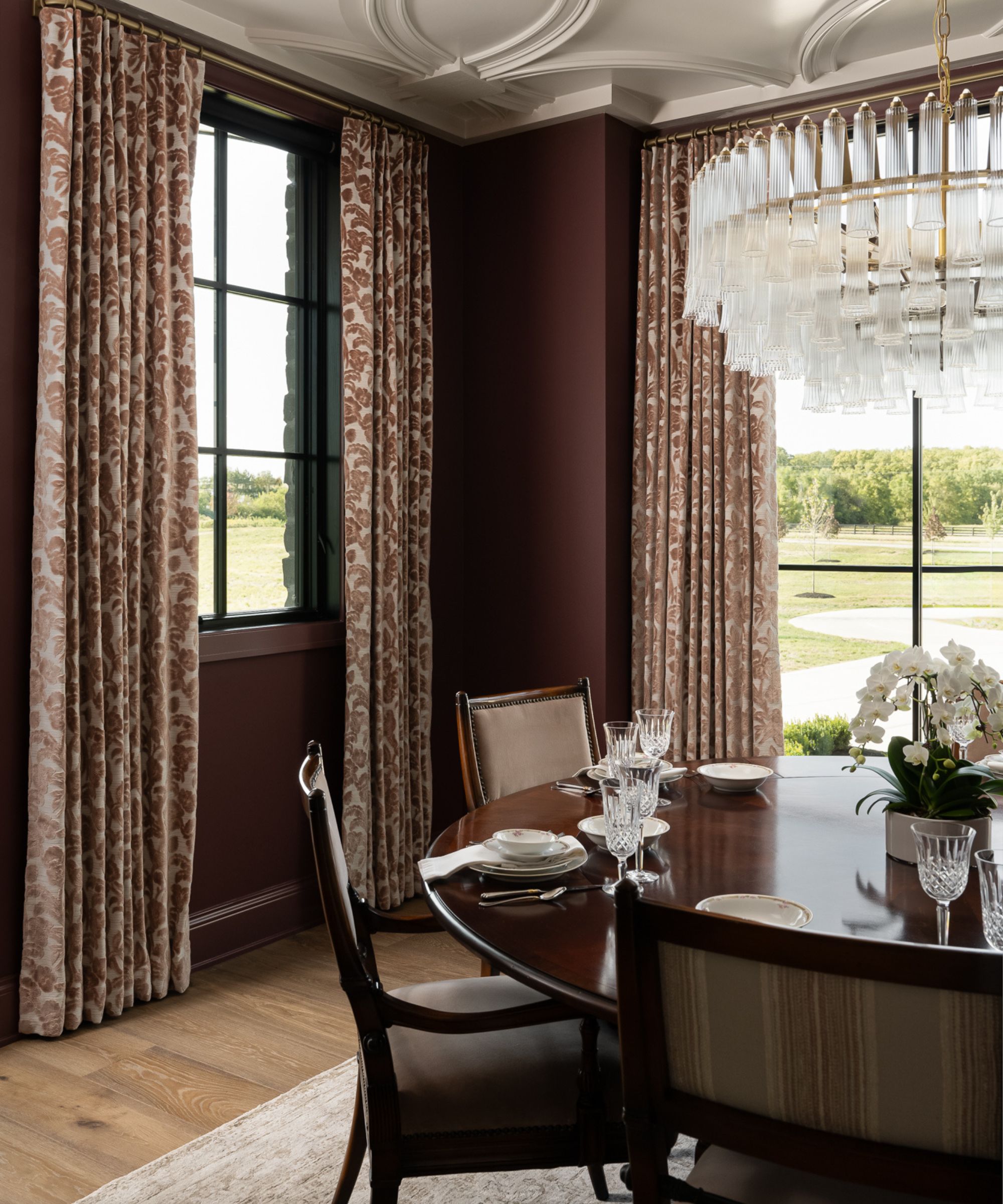 A formal dining room with deep burgundy walls, a dark wood table set with fine china, patterned curtains, and a large tiered glass chandelier.