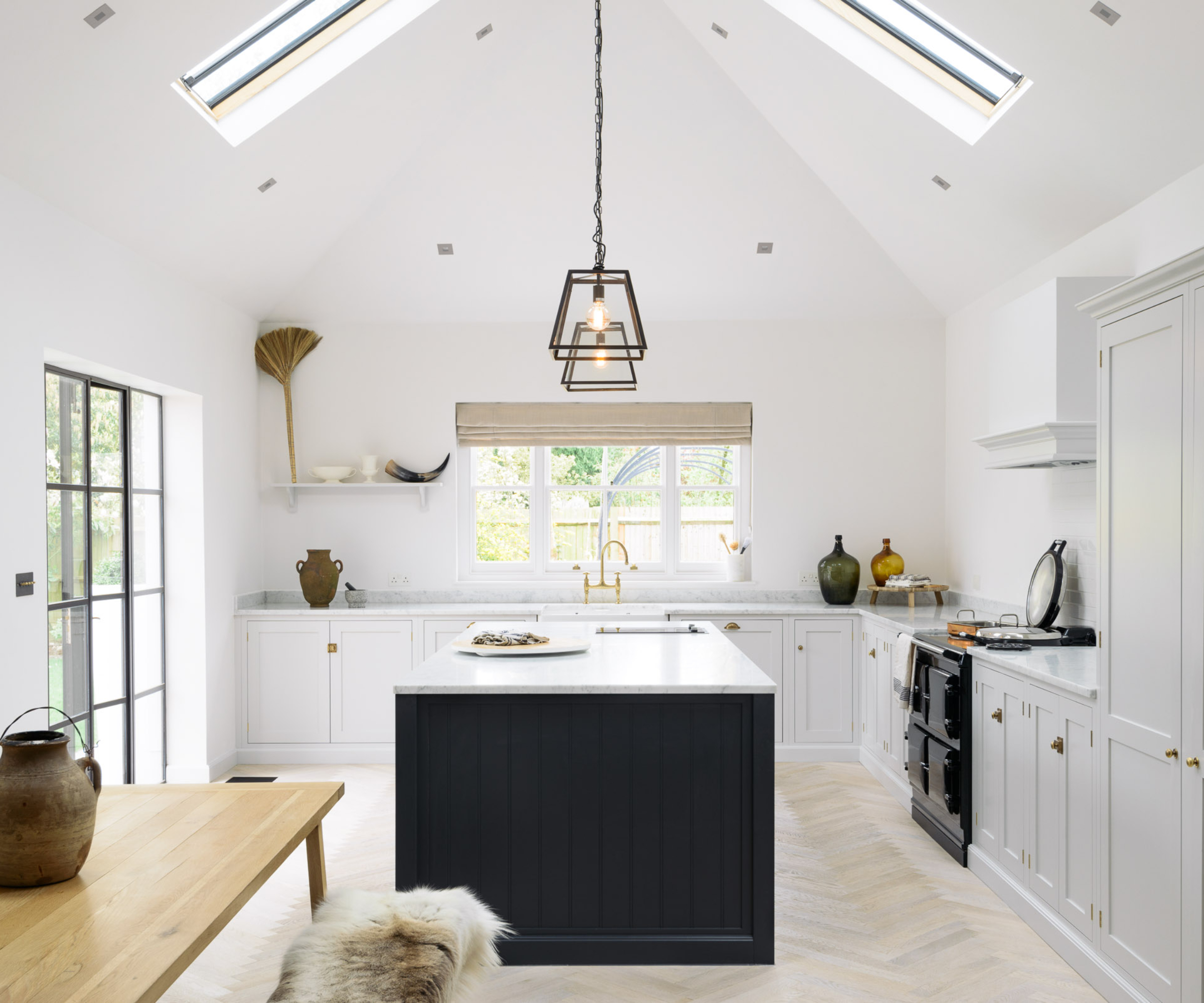 large kitchen with pitched roof and skylights, white kitchen units, navy based kitchen island light wood kitchen floor, light wood dining table, pendant lighting