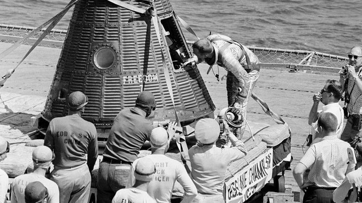 black and white photo of an astronaut inspecting his small space capsule on the deck of a ship at sea