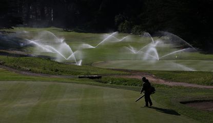 Sprinklers go off in the background whilst a greenkeeper brushes leaves away