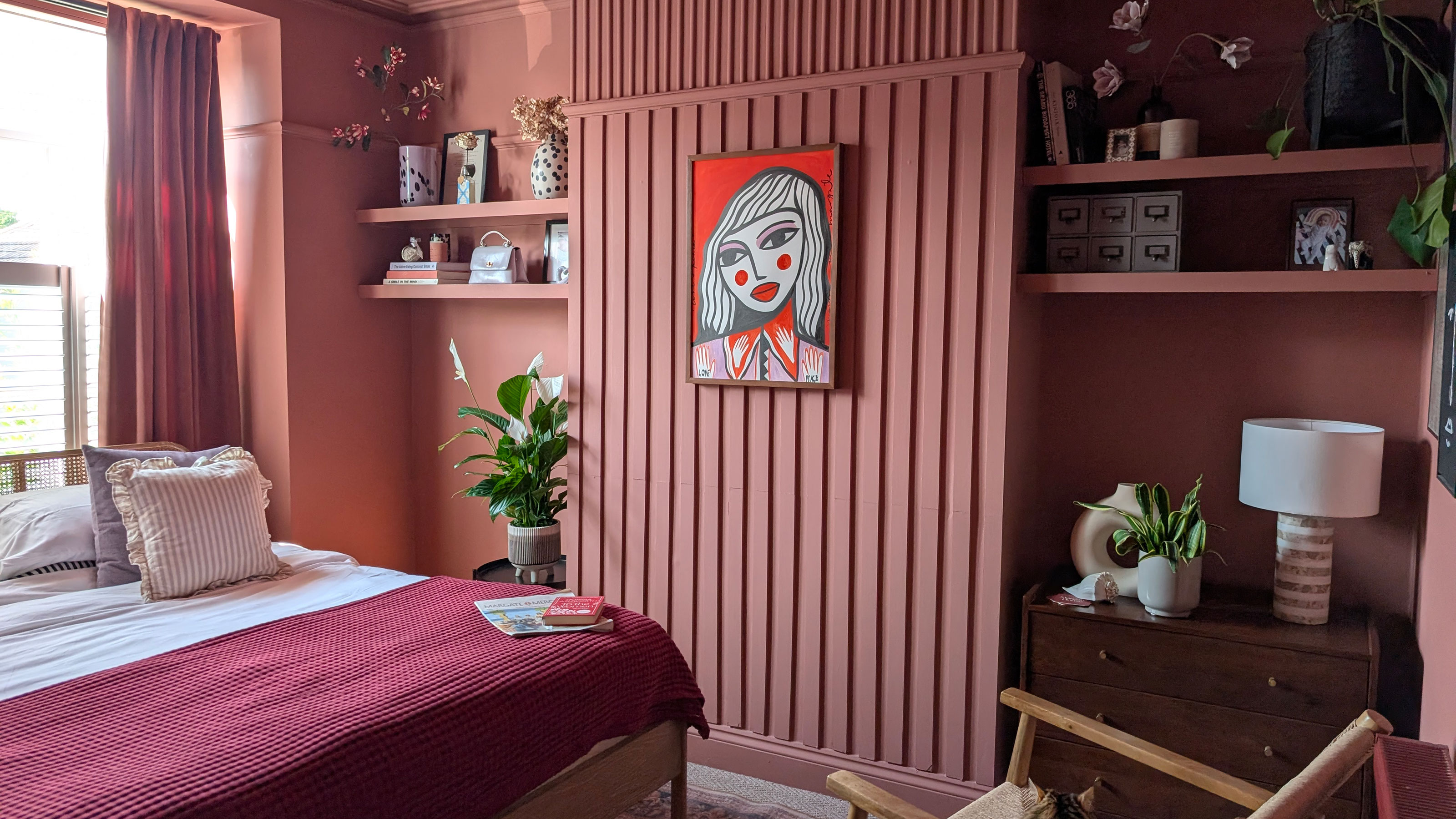 Pink bedroom with wall panelling, artwork of woman's face on wall, floating shelves and red throw on bed