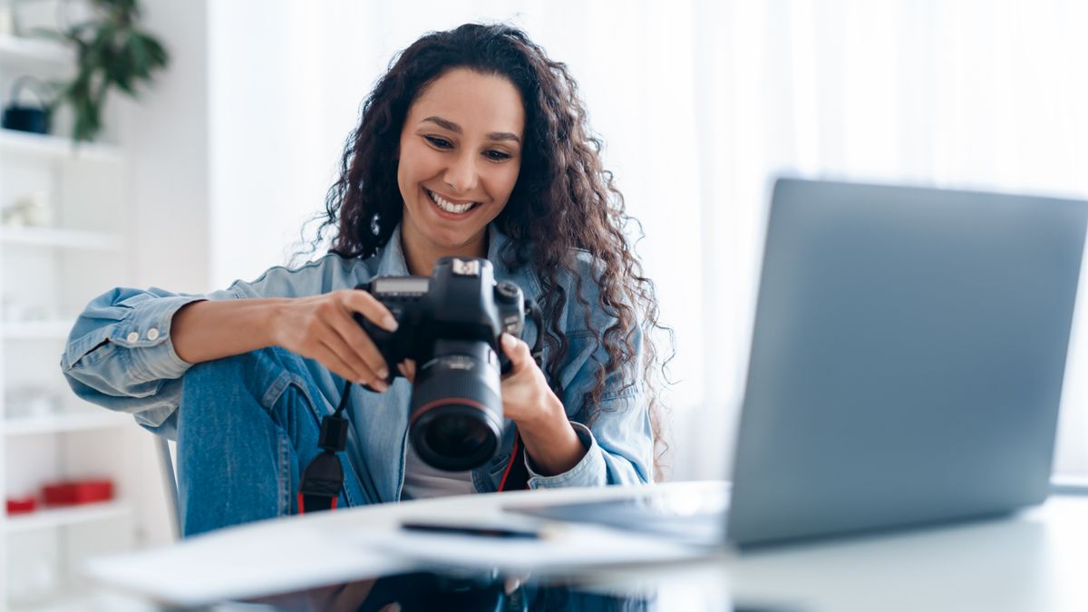 Joyful Professional Photographer Lady Holding Camera Using Laptop At Workplace