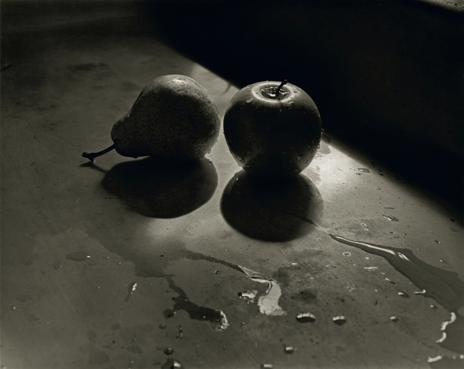 A black and white close-up of a pear and an apple resting on a wet surface, casting dark shadows beneath them.
