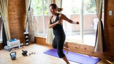 A woman workouts out energetically at home. She is jumping to one side, balancing on one leg with her arms held in the air. Behind her we see an exercise mat, abs roller, dumbbells and kettlebell, along with a large window framed by curtains.