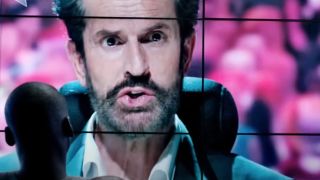 Rupert Everett on a screen, looking serious with the back of head watching the him in Black Mirror