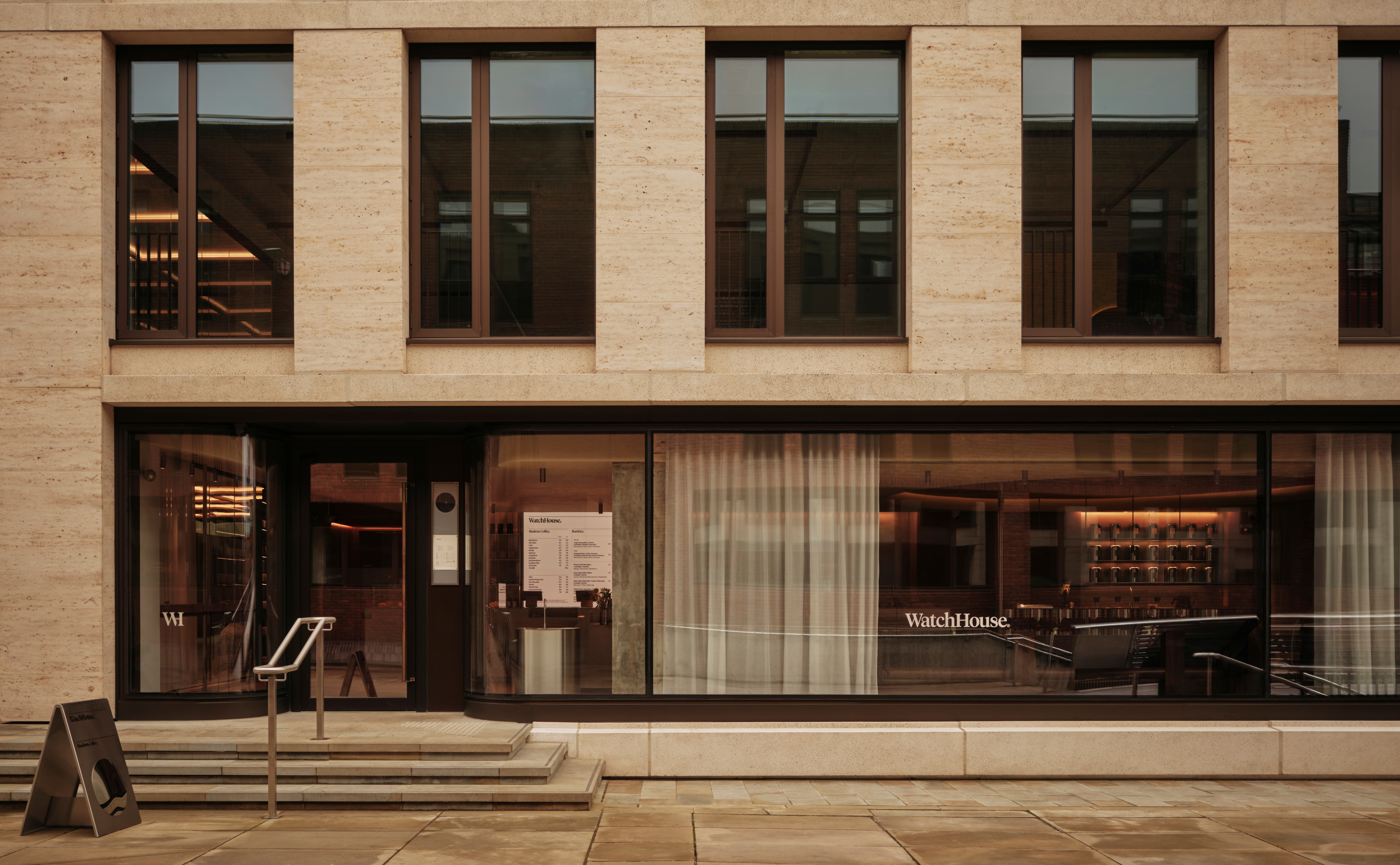 The wood, chrome, and white textile-clad interiors of a cafe boost chocolate leather mid-century furniture, dramatic ceiling lighting, and plenty of natural light coming through floor-to-ceiling windows.