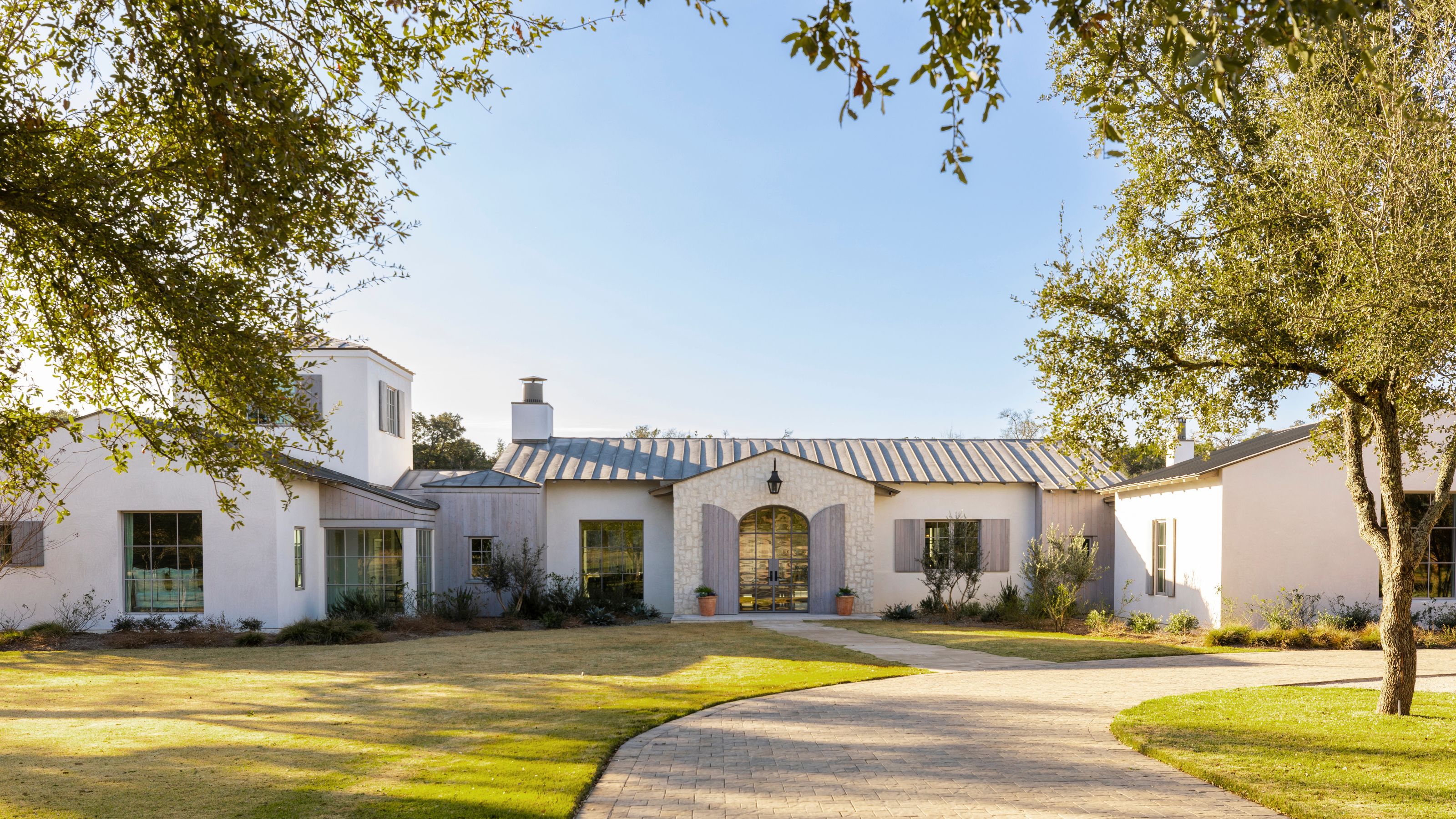 a large Texan farmhouse with a low white exterior, window shutters, large lawn and driveawy and trees