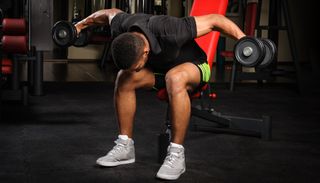 Man sits on edge of weights bench performing reverse fly with dumbbells