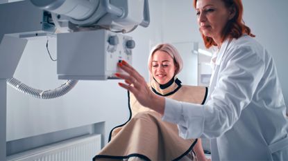 Smiling, cheerful young woman is wearing a protective lead apron (patient). She is talking with female doctor who is turning on x-ray machine for foot scanning in a modern hospital.