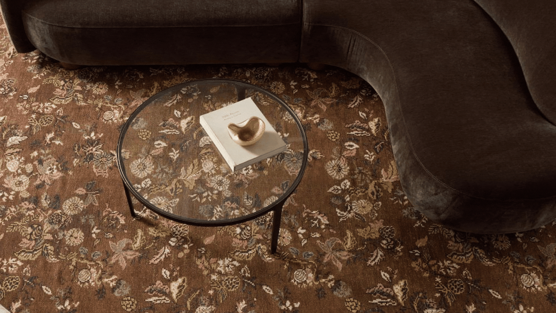 A GIF of two images showing either side of a brown patterned, reversible rug from above. There is a brown, curved sofa and a round glass coffee table on the rug.