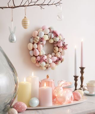 A pastel colored egg wreath hanging on a white wall with a marble side table underneath it with white, pink, and white candles, eggs and rabbit decorations on it, with a branch of egg and rabbit decorations above it