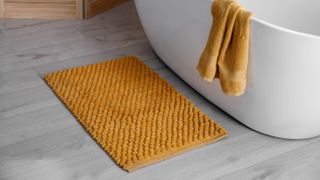 A yellow bath mat on the floor nect to a bathtub with a towel thrown over it