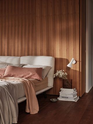 small modern bedroom with wooden wall, off white modern bed, wall light, hardwood floor, pile of books with vase on top, neutral and earthy bedding and blankets