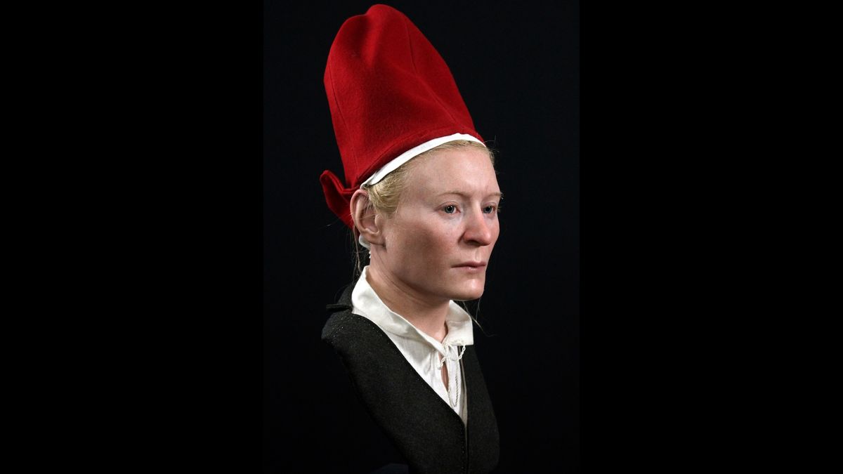 We see the reconstruction of a woman&#039;s face and upper torso. She has pale skin, blonde hair and a solemn expression. She wears a tall bright red hat, a gray jacket and a white blouse with a collar. 