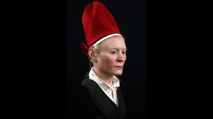 We see the reconstruction of a woman's face and upper torso. She has pale skin, blonde hair and a solemn expression. She wears a tall bright red hat, a gray jacket and a white blouse with a collar.