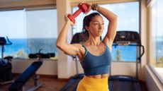 A woman in a tank top and leggings performs a dumbbell halo in a gym. She is standing, arms slightly bent, a dumbbell held in her hands over the top right side of her head. 