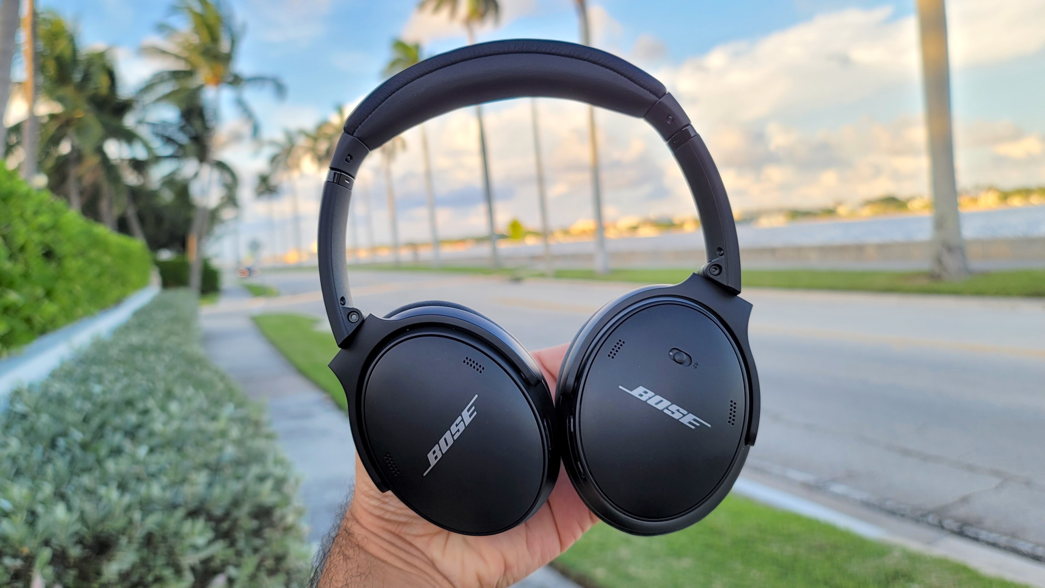 The Bose QuietComfort 45 headphones being held aloft against a backdrop of a coastal street with palm trees