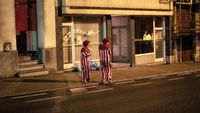 Two clowns stand talking on a night-time pavement outside a shuttered baby goods shop, lit by the shop's glowing window display.