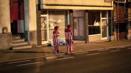 Two clowns stand talking on a night-time pavement outside a shuttered baby goods shop, lit by the shop's glowing window display.