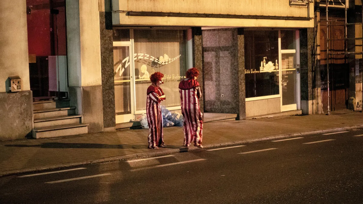 Two clowns stand talking on a night-time pavement outside a shuttered baby goods shop, lit by the shop's glowing window display.