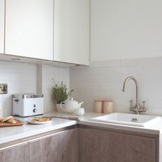 White kitchen with sink area and toaster, teapot, plant and sliced bread on chopping board on white worktop