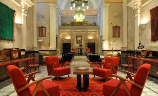 Common area of the L’Iglesia Hotel — El Jadida, Morocco, with red upholstered chair and carpets, wall hangings and wooden furniture