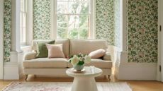 A traditional green living room with a white sofa in a window alcove. Green floral wallpaper and white moulding on the walls. A small round white ceramic table.