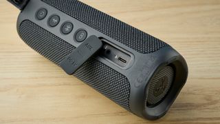 a close up picture of the aux jack a black oblong-shaped bluetooth speaker with mesh covering and a carry strap photographed on a beige table in front of a navy wall