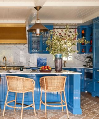 Blue kitchen with an island, terracotta floor, marble worktops and splashback and brass pendant lights