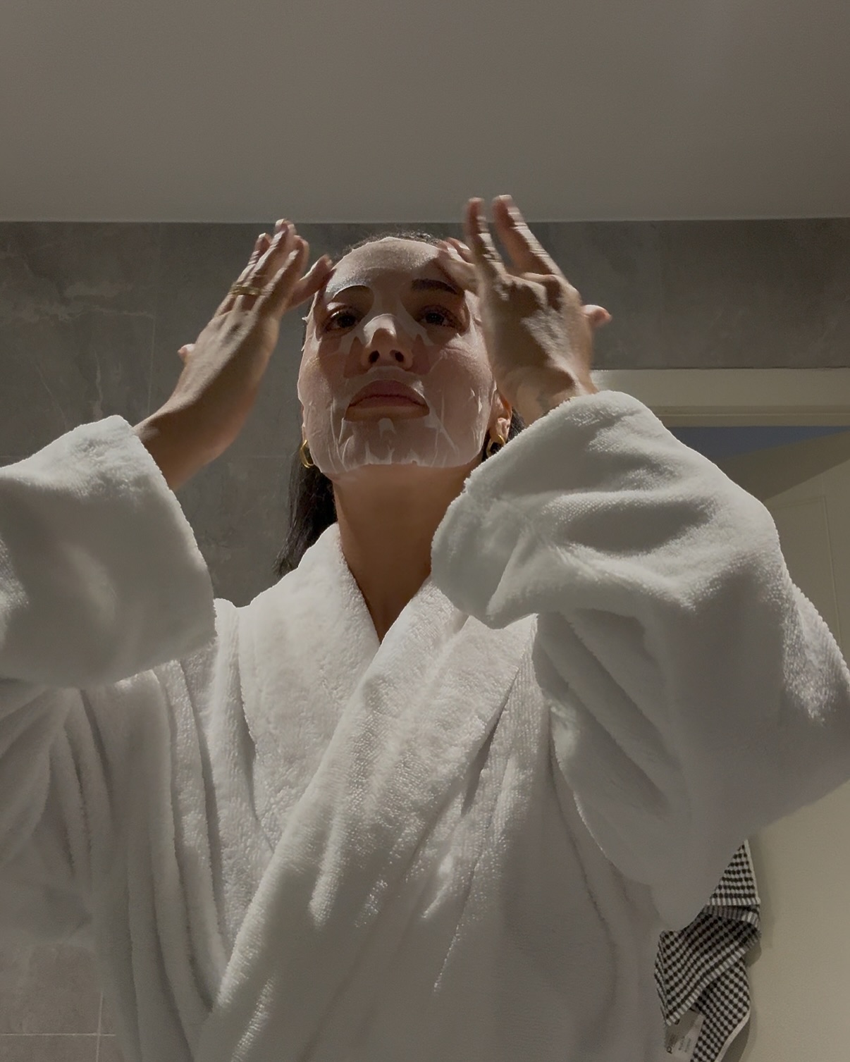 Photo of a woman putting on a sheet mask in a white robe