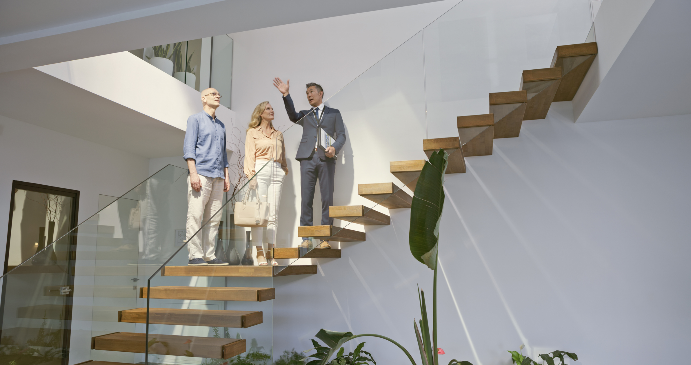 Male estate agent showing modern luxury house to mature couple while standing on staircase in living room.