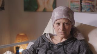 Lucy Lowe as Jodie's mum Rachel in a hospital bed in her living room, wearing a grey hoodie and pale lilac headscarf with white stars, and looking sad and troubled.