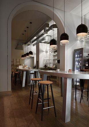 View of the bar in the restaurant with large white countertops, high ceilings with long bar lights in black, and metal and wood bar stools.