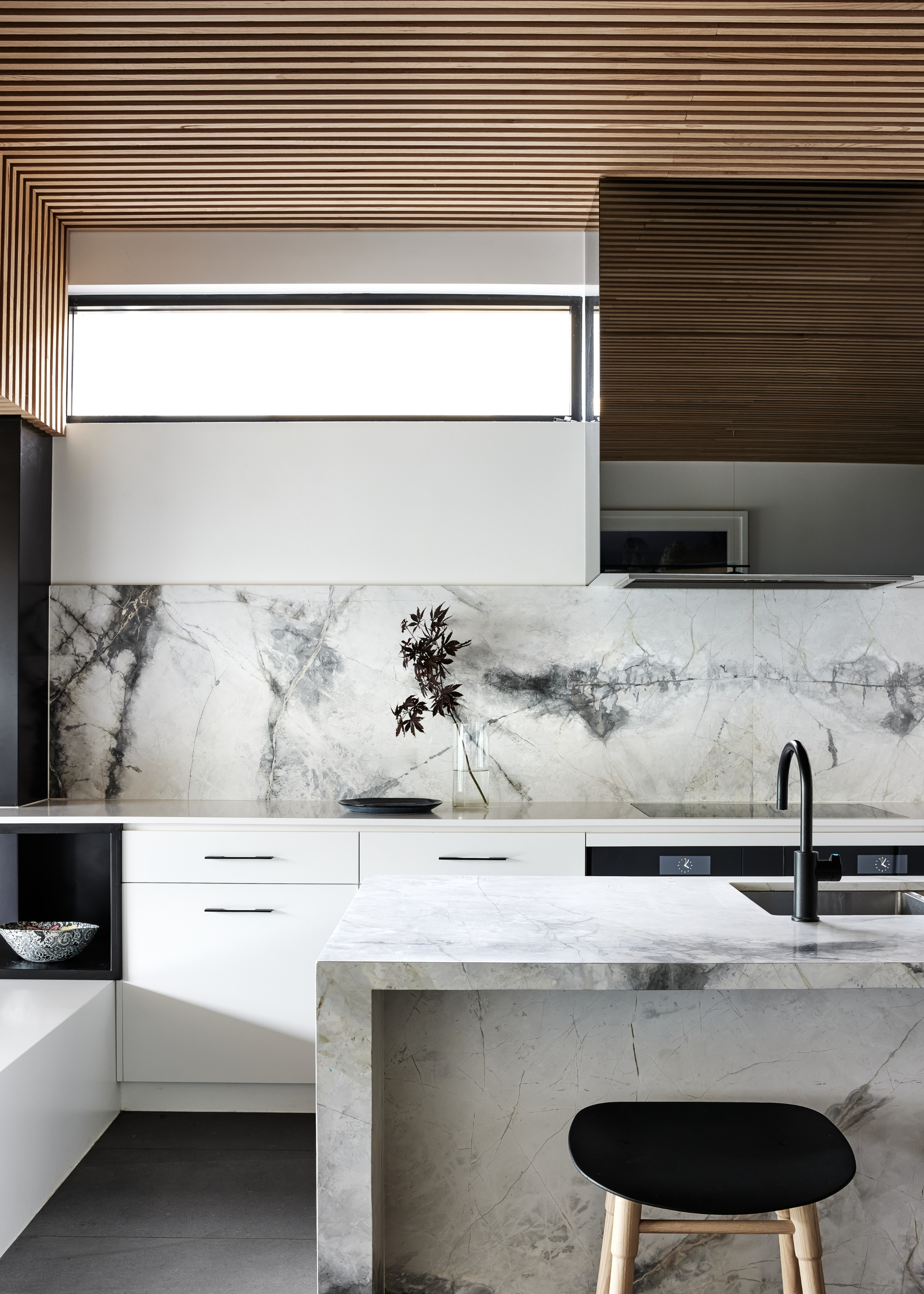 A kitchen with an island in the middle and a bar stool beside it. On the island is a sink and black colored tap. There is also a view of the rest of the kitchen and ceiling that has built-in wood panelling.