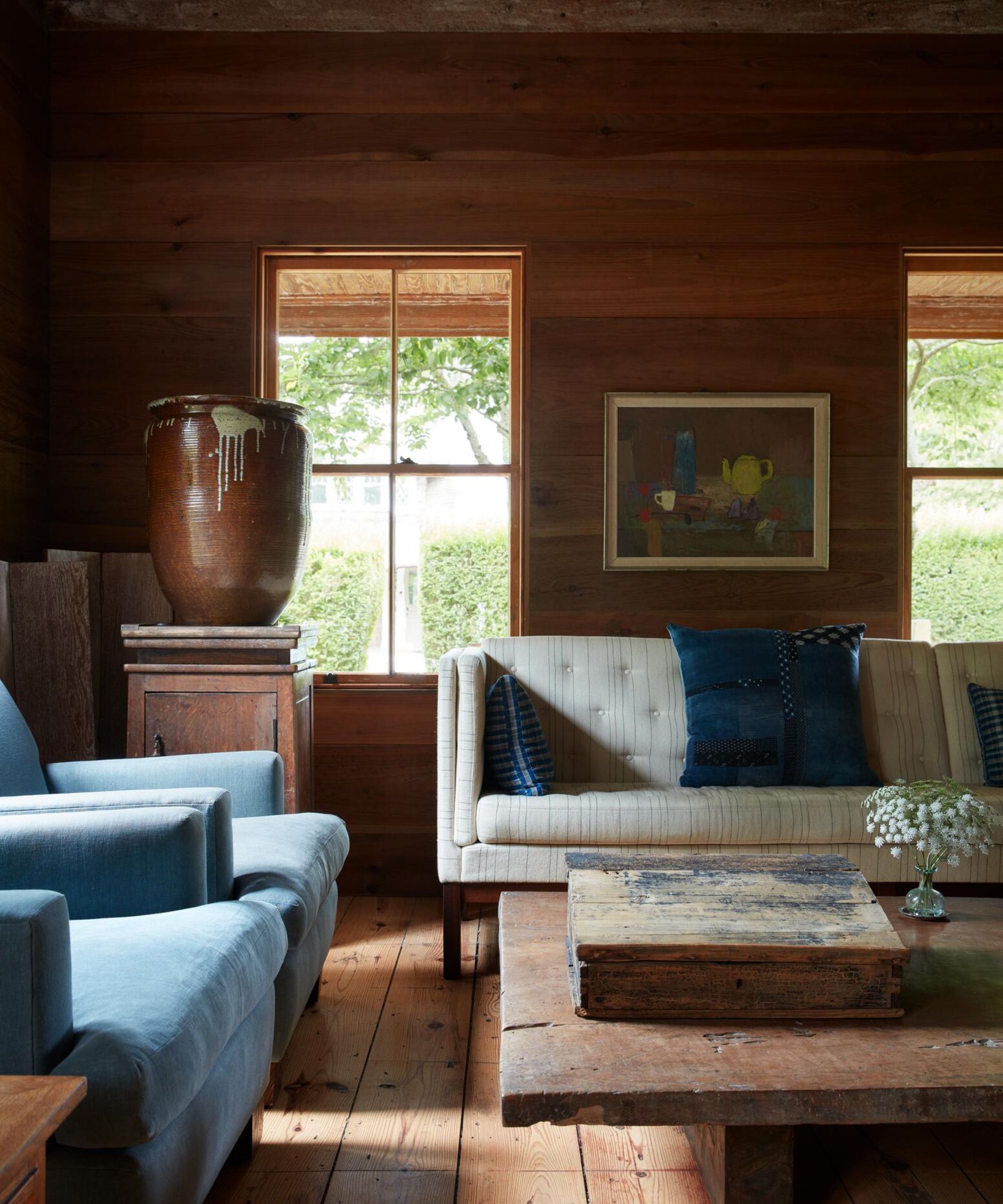 A sunlit living room with wood-paneled walls. It features a neutral tufted sofa with blue patterned pillows, two light blue armchairs, and a low, chunky wooden coffee table. A large ceramic jug sits on a pedestal by the window.