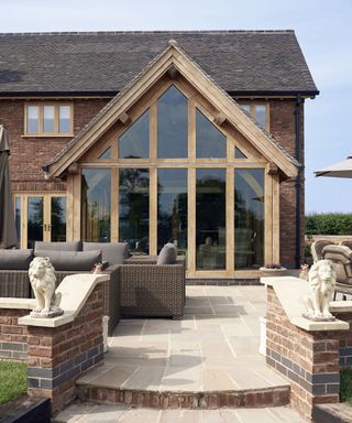 A large wooden extention with glass windows and a pitched roof on the end of a brick home. Paved patio with rattan garden furniture in front.