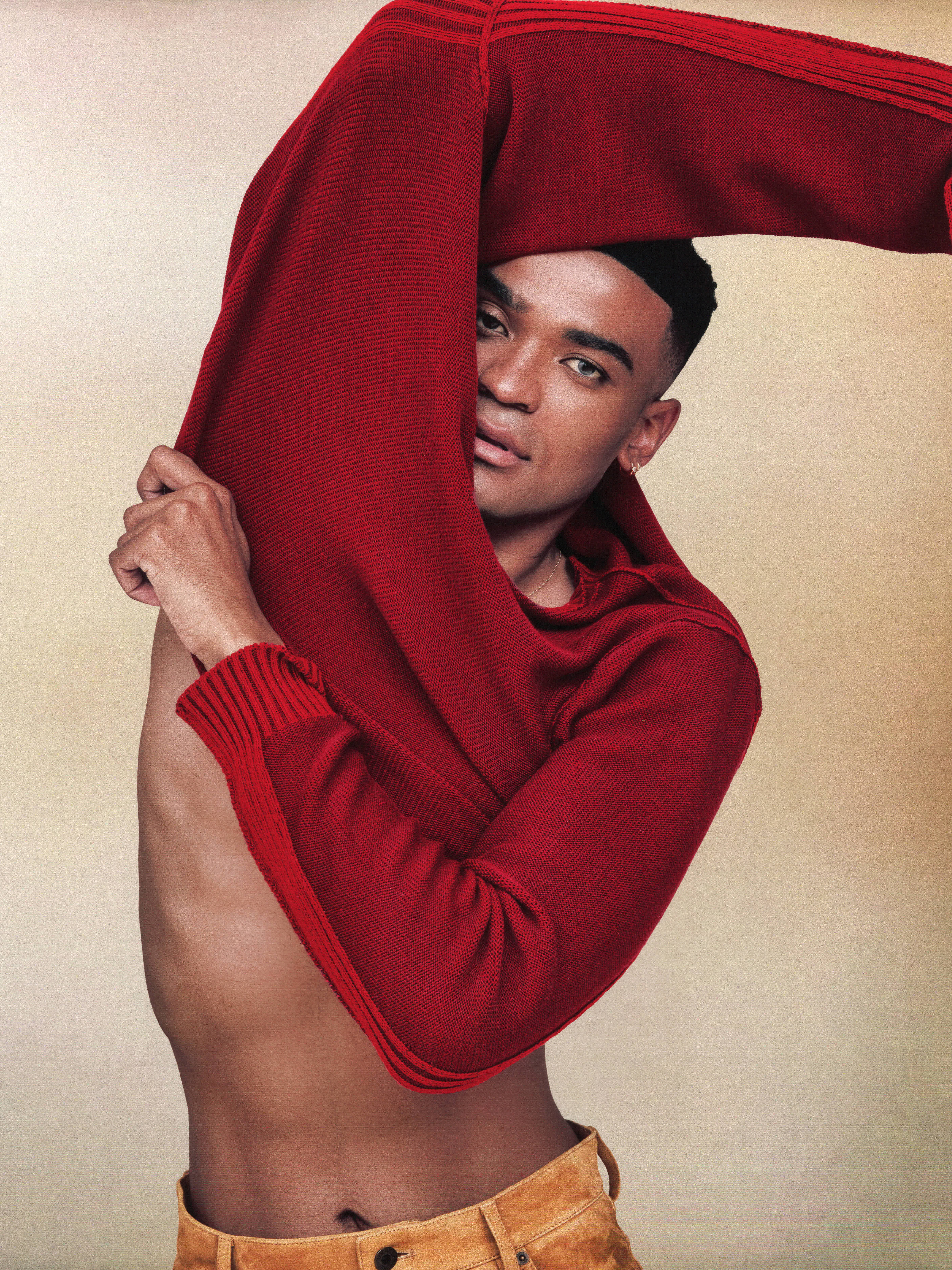 Owen Thiele poses wearing a red sweater that reveals his stomach as he lifts his arms over his head