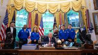 four people in blue flight jackets stand behind an old man at a desk in a grotesquely decorated office